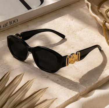 Black luxe sunglasses with gold detailing displayed on a beige stone surface