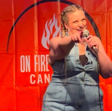 Female stand-up comedian performing on stage with a microphone in front of an On Fire comedy backdrop.