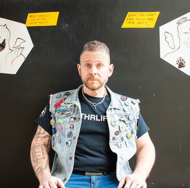 man in goatee with denim vest covered in pins with cartoon angel and demon on black background