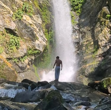A man stands on rocks looking at a powerful tropical waterfall cascading down a lush cliffside.