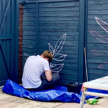 Muralist sits on floor painting graphic plant on grey wooden garden fence.