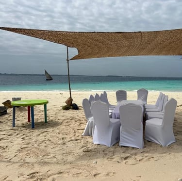 Shaded dining setup with white chairs and table on Nakupenda sandbank, Zanzibar, with beach views and a small local dhow
