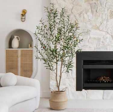 Tall artificial olive tree in a woven basket next to a modern fireplace and white stone wall.