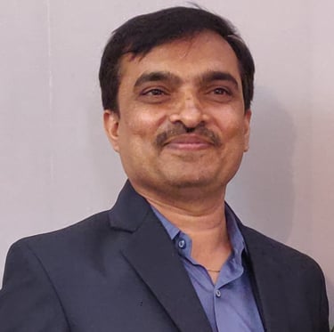 Portrait of a man wearing a dark suit jacket and blue dress shirt, with short black hair and a mustache, smiling in front of