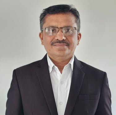 Portrait of a man wearing a dark suit jacket and white dress shirt, with glasses, short black hair, and a mustache, standing
