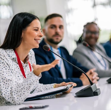 A smiling female speaker using a gooseneck conference microphone during a formal panel discussion, seated alongside two other
