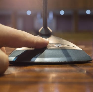 A close-up image of a person's finger pressing the power button on a gooseneck conference microphone base placed on a wooden 