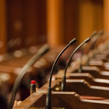 A row of gooseneck microphones installed on individual wooden podiums inside a formal conference or parliamentary setting, wi