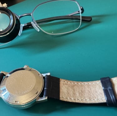 Vintage watch back, jeweler's loupe, and eyeglasses on a green surface for watch repair.