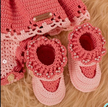 Handmade pink crochet baby booties and dress with pearl bead accents on a soft faux fur background.