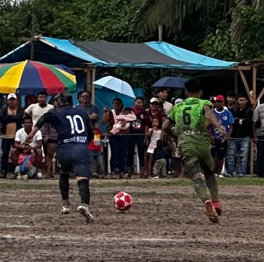 Soccer players competing on a muddy field during a local football match with spectators in the background.