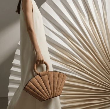 Woman in white dress carrying a beige cork fan-shaped handbag against a pleated background.