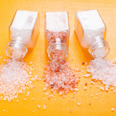Three glass jars spilling different types of Himalayan salt granules on a bright orange surface