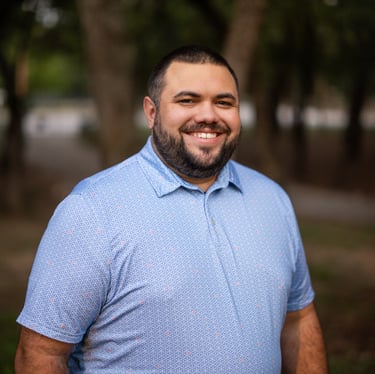 Photo of Jake Gigl smiling outdoors, wearing a blue patterned polo shirt, with trees in the background.