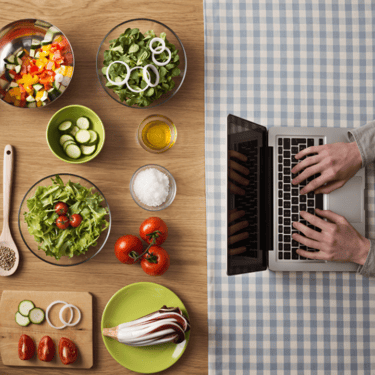 Person typing on laptop at other end of table that displays many dishes/ingredients