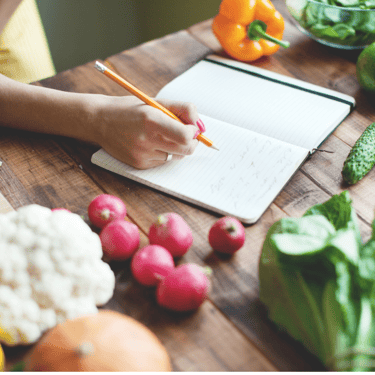 Photo of a person writing in a journal on a table that also has colorful ingredients on it
