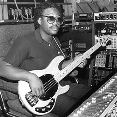 Black and white photo of Bernard Edwards holding a Music Man bass. He's sitting at a mixing console.
