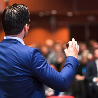 A keynote speaker in a suit presenting to a large audience at a conference.