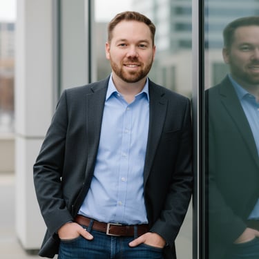 Man in blazer and jeans leaning against glass wall with city buildings in background.