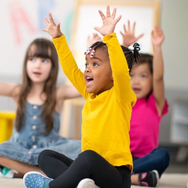 Photo of children in classroom with arms in the air.