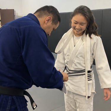 A Brazilian Jiu-Jitsu instructor awards a grey belt to a smiling female student in a BJJ gi.