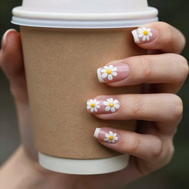 Close-up of hand-painted press-on nails featuring tiny daisy patterns and soft blush pink accents, held against a warm coffee cup.