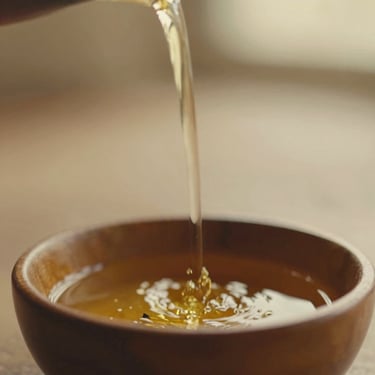 Close-up of warm herbal oil being poured into a small wooden bowl, soft diffused golden-hour light, earthy muted tones, shallow depth of field, 35mm film