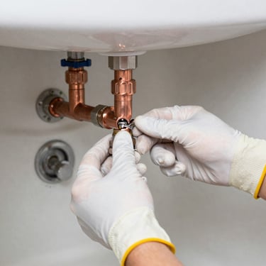 Close-up of a technician's hands in protective gloves repairing a copper pipe under a sink, bright daylight.