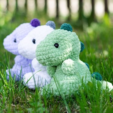 Tres dinosaurios amigurumi de crochet de color verde, blanco y morado sobre el pasto