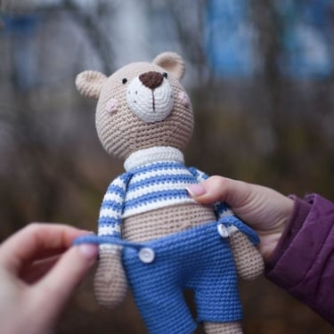 Osito amigurumi tejido a crochet hecho a mano, con suéter a rayas y overol azul.