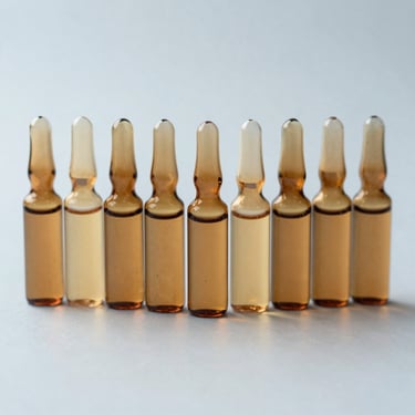 Overhead close-up of small amber glass remedy vials arranged in a row on a plain pale surface, soft studio light, no labels visible, 1:1 scale