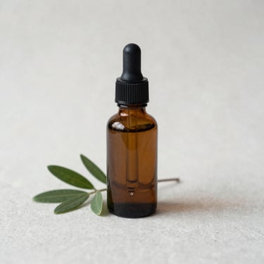 Close-up of a small dark glass dropper bottle on a plain pale linen surface, north-facing soft daylight, botanical sprig beside it, 1:1 scale, no people