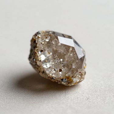 Single raw crystal specimen placed on a plain pale linen surface, soft north-facing studio light illuminating its facets, no props, extreme close-up showing mineral texture and colour
