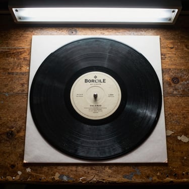 7-inch vinyl single out of its sleeve on a worn wooden table, venue fluorescent light above, label text visible, shot from directly above at close range