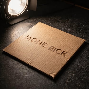 Album sleeve flat on a dark stage floor, warm side-lighting from a practical stage lamp, textured cardboard sleeve with rough-cut lettering, shot tight from above at a slight angle
