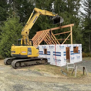 Yellow Komatsu excavator lifting its bucket over a roof frame on a building wrapped in Tyvek HomeWrap and RONA branding.