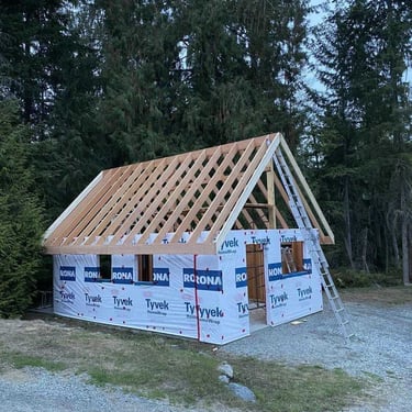 Small building under construction with a wooden gabled roof frame and Tyvek HomeWrap siding on a gravel pad in a wooded area.
