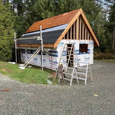 Wooden structure under construction in a wooded area with a gabled roof, Tyvek HomeWrap siding, and ladders on a gravel pad.