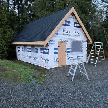 Small building under construction in a wooded area with a steep gabled roof, Tyvek HomeWrap, and ladders on gravel.