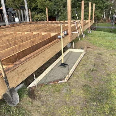 Wooden deck frame under construction with concrete form, shovel, and posts in a grassy yard.