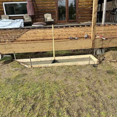 Wooden deck construction attached to a log cabin with concrete footing and scattered tools.