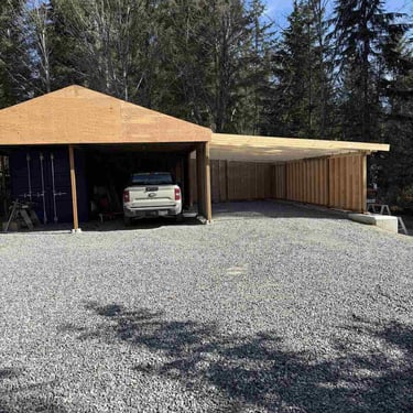 Open-front wooden carport or shed under construction with a blue shipping container and pickup truck on gravel.