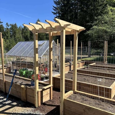 Wooden garden arbor over a gravel path with raised beds, greenhouse, and evergreen trees in the background.