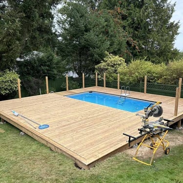 Wooden deck under construction around a blue pool with ladder and miter saw.