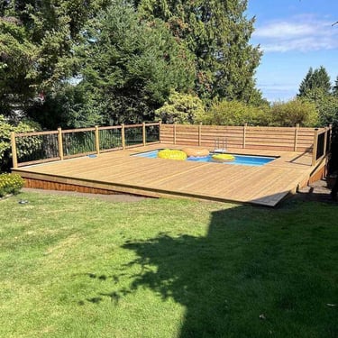Above-ground pool on a wooden deck with inflatable rings, privacy fencing, and evergreen trees under a clear sky.