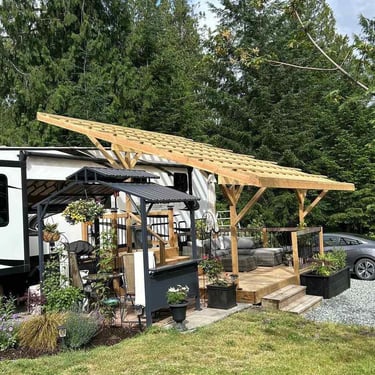 White RV and timber-frame roof over a deck with gazebo, outdoor furniture, planters, and evergreen trees.