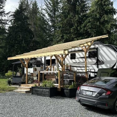 White “REFLECTION” fifth-wheel RV with wooden pergola deck, planters, and a grey sedan on gravel beside evergreen trees.