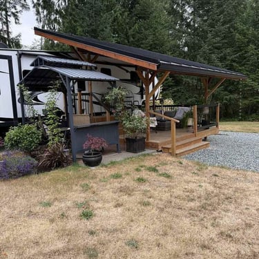White RV with custom wooden deck, slanted metal roof, gazebo bar, potted plants, and evergreen trees.