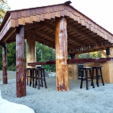 Rustic log outdoor bar with tiered shingle roof, wooden counter, stools, bottles, sandy ground, and a white dog.