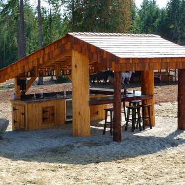 Rustic wooden outdoor bar with stools, bottle shelves, and horses in the background.